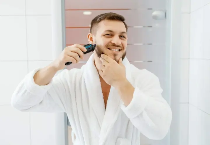 Mężczyzna w białym szlafroku goli zarost trymerem.