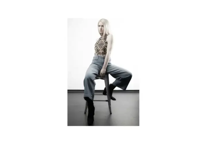 Blond modelka w luźnych jeansach i wzorzystym topie siedzi na stołku.