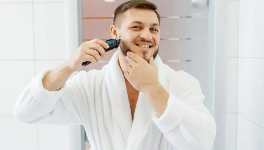 Mężczyzna w białym szlafroku goli zarost trymerem.