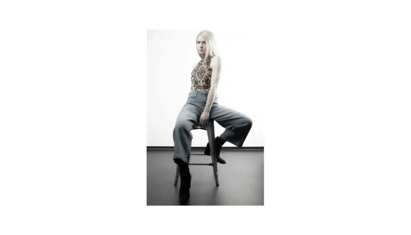 Blond modelka w luźnych jeansach i wzorzystym topie siedzi na stołku.